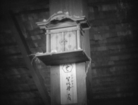 A small shrine is attached to a wooden pillar, above a calligraphic sign.
