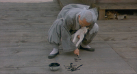A squatting man paints calligraphy on floorboards with a white cat's tail, seen from the front.