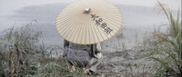 Black calligraphy on a paper umbrella, held by a person crouching alongside water in the mud.