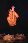 A half-naked man kneels in a pile of scattered paper against a black background. His left hand is raised just above his face, slightly covering it, while the right hand holds the left wrist.