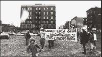 Black and white photograph of group standing in rubble strewn lot holding up a banner.