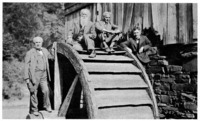 The four friends on vacation. Left, Edison standing on hub of old water wheel, Burroughs and Ford on the wheel, and Firestone to the right of it.