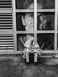 In this black and white photograph, a person wearing pants and sneakers, huddled on the street and wrapped in a blanket so that the entire upper body is concealed, sits on a ledge outside a large ground floor window on an otherwise deserted city street. Behind the person, in the windows of the building, is an oversized poster featuring a photographic image of a man’s face with a slight scowl.