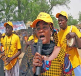 A woman speaks into a microphone during an outdoor demonstration. She and the two men behind her wear yellow baseball caps.