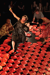 Fig. 8. Raised stage shot from above, covered in red plastic plates. Black woman and two others placing the plates; white woman with gag fake eyeglasses slips on a plate.