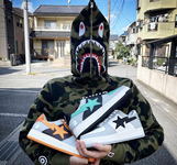 A man stands in the street wearing a camouflage print sweatshirt with a shark hood that zips over his face. He is holding three pairs of gray and white sneakers with a shooting star printed across the outside. One shooting star is orange, one turquoise, and one black.