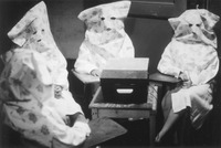 Four women sit in a half circle around a small table with a box-­like machine on it. They are wearing oversize white lab coats and large hoods over their heads, apparently made from pillowcases bearing a flower design. Small eye and mouth holes are cut into the hoods.