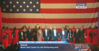 Ben Carson, his family, and two singing groups, Veritas and Selected of God, appear singing on stage in front of a very large American flag.