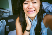A photographic close up of a young Asian woman in a sleeveless blue-and-white shirt a crying with closed eyes. One hand is on her neck.