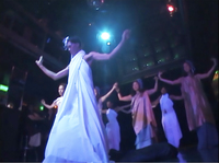 Screen capture of Akuna, a tall brown figure in a long white wrap, in midmovement with arms out in front of other dancers on a stage.
