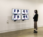 Photo: a person with headphones on views a video installation featuring four screens showing very similar images of Stephanie Dinkins, a Black woman with shoulder-length locs, wearing a white T-shirt and colorful scarf mimicking the actions of and talking to Bina48, a Black female robot with shoulder-length, light-brown hair wearing a white T-shirt and colorful scarf.