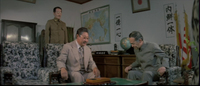 Men converse in an office with black and white calligraphy on a desk placard and banners and a map on the wall.