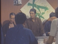 A group of people surround a table, with a large green calligraphic character on the wall behind them.