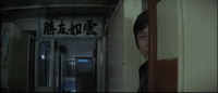 Image of a man peeking down a hall from behind a wall. In the distance down the other side of the hall, a large banner with black calligraphy hangs.