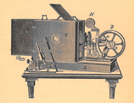 Line drawing of a prototype movie camera on a table. Film reel, hand crank, lightbox, and other parts are labeled with letters.