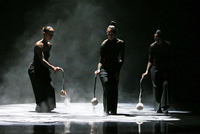 Color photograph of three female performers dressed in black, shaking out white dust from their poi on to a darkened stage.