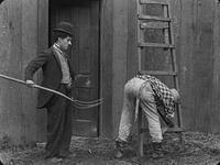 Fig. 34. Charlie Chaplin as the Tramp character, dressed in baggy clothing and bowler hat, holds a pitchfork near the posterior of a man who is bending over to tend to a task outside a barn, near a ladder.