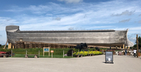 This is a photograph of the Ark at Ark Encounter. It is taken from the position of visitors approaching the ship from a distance and shows one entire side of the gray timber-made Ark. The Ark is framed by the sky above it and decorative grass landscaping below.