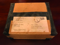 Color photograph of a cardboard box with a faded shipping label identifying the contents as US flag belonging to Larry R Cooper.