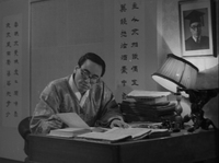 A man sits in his office in front of banners with black calligraphy printed on them.