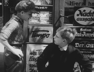 Two children sitting in front of a newspaper kiosk, talking.