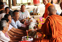 Three Cambodian Buddhist monks in orange robes and shaved heads give blessings to a group of smiling villagers at a long, low table covered with candles, platters of fruit, and various other offerings.