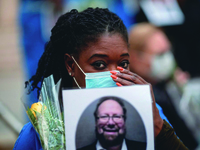 A middle-aged Black woman wearing a protective mask gazes toward the viewer. With one hand, she holds her mask in place. With the other, she holds a bouquet of yellow roses and a posed photograph of a bearded white man in her other hand. The man in the photograph—presumably now deceased—is smiling.