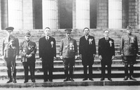 This black-­and-­white photo shows seven men standing in front of a neoclassical building with Greek columns and stairs, all wearing ribbons and medals. Ba Maw, Zhang Jinghui, Tōjō Hideki, and Subhas Chandra Bose are in military uniform, while Wang Jingwei, Wan Waithayakon, and José Laurel are in dark tuxedoes. Wang Jingwei, the tallest, is standing to the left of Tōjō, with close-­cropped hair and looking slightly to the left. Zhang Jingwei’s hands are folded together in front of his belly, looking a little unnatural. Ba Maw and Tōjō are holding sabers in their left hand.