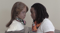 Photo: On the left is Bina48, a female robot with dark-brown rubber skin with shoulder-length, light-brown hair wearing a white T-shirt and colorful scarf. On the right is artist Stephanie Dinkins, a dark-brown woman with shoulder-length locs, wearing a white T-shirt and colorful scarf. There is a light-gray background.