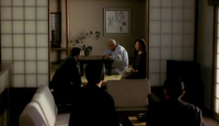 Men gather outside a room where a woman and elderly man sit. A poster hangs on the wall with black calligraphic writing.