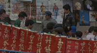 People walk through a narrow street, with posters in the background and red and gold calligraphic banners in the foreground.