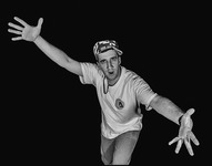 Solo performer leaning forward with arms extended and fingers splayed.  He is wearing a hat, beaded necklace, t-­shirt and jeans and looking into the camera as he speaks.