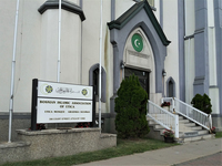 A front view photograph of the building of the Bosnian Islamic Center of Utica, NY taken in August of 2021. High on a hill, the building is located at 306 Court Street, in the center of the city and right above Utica City Hall. It is built as an Anglican style church with tall steeples that is now to be seen (but not really used as a minaret), and was formerly housing a Trinity United Methodist Church. The building was in disrepair and was awarded to a Bosnian Muslim Community by the city for much smaller price than its actual worth, with certain conditions to preserve the structural integrity of the building, while allowing some visual modifications to the façade. In the image we can see the introduction of green paint with a white crescent moon and star, as well as a sign indicating that building is now “Bosnian” Islamic center, which is uncommon for the US Islamic places of worship. Most of the Islamic Centers in the US will not indicate any national associations, no matter how ethnically homogenous congregations might be, with the exception of some Turkish Islamic Centers. Furthermore, it is located just down the street from the “West Bosnian” dedicated Islamic Center. Both centers will, on special occasions, host three flags: Bosnian, American, and the green flag of the official religious hierarchy of IZBiH.