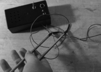 Black and white photo, a close up of two metal sticks in a person’s hand. These are connected by wires to a small black box with two switches.