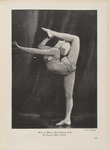 Black-and-white photograph of a dancer in front of a black curtain wearing a beaded bikini in a seemingly improbable feat: she holds a pose on one foot, while the other leg bends back so that her leg wraps behind her body and up over her head. She arches her back and situates her head behind her knee, so her foot is ahead of her face and her calf touches her nose. She holds the raised leg with one hand and stretches the other arm outward.
