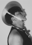 Black-and-white photograph of Marianne Brandt, acting master of the metal workshop, dressed for the Metallic Festival in her own creations. Shown here in a self-portrait, she wears a metal plate on her head with a strap fastener like a hat, and a ball hangs from a large, thick metal ring on her neck like a necklace. She stands facing our right with her head tilted slightly upward.