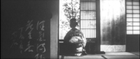 White calligraphy on a black background decorates a fusama sliding door. It fills the left hand side of the frame. A woman sits in the middle of the frame in the background, looking out a window.