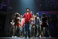 The character Usnavi center stage in a red shirt, cap, and blue jeans. The ensemble crowds around him in front of the bodega that is central to the musical.