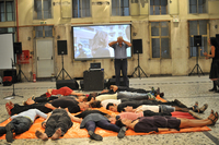 One man stands in front of a projection screen and covers his eyes with his hands. In front of him, a group of dancers are lying down on a large orange tarp.