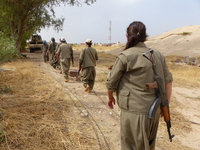 YJA Star guerrillas at the frontline against Daesh, 2015, Kirkuk, Iraq. A line of six women walk down a dirt path behind an armored truck in a dry territory, marching toward their training area. They are wearing camouflage uniforms, different-­colored caps, and Mekaps. All in the row are armed, each carrying an AK-­47.