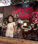 Pink graffiti paint on a highly graffitied wall in Beirut reads “Queers for Marx.” All the other written graffiti is in Arabic. On the bottom left of the wall is a graffitied picture of a child with black hair and brown skin in a white dress.