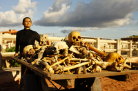 A woman pushes a handcart full of human bones on a red dirt road. She has cropped black hair and is dressed in a long black gown with turtleneck. The sun shines upon her and the bones. Behind her is a blue sky and heavy clouds. There is also a white rail that indicates this may be a bridge