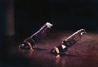 Two dancers balance their bodies at an angle with faces on the floor and pointing their legs in parallel into the air. One is wearing a baseball cap and the other a black hoodie. They both wear Adidas-­style track pants with white stripes down the side.
