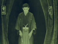 Green-toned image of a performer standing between two curtains, looking down. The performer wears a special outfit and holds a fan; the curtains have calligraphic emblems on them and are parting to make way for the performer.