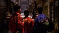 A trio stands on a street with yellow calligraphy and white English text printed on the back of their jackets. Banners and store signs have red and white calligraphy printed on them.