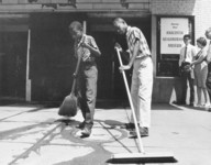 Black and white photo of two youths sweeping a sidewalk in front of a building. In the background are three adults and sign is visible which reads, “Anacostia Neighborhood Museum.”