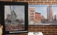 Digital photograph of one part of an exhibition in which a hand holds up a transparent viewer that shows the Statue of Liberty as Our Lady of Guadlupe. Through this viewer, behind it, you can see a large-scale panoramic image of the neighborhood on the wall.