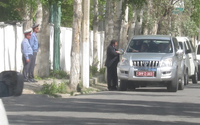 One man in a suit who belongs to the Tajik security services approaches a diplomatic in a black SUV with a red license plate. The man is talking to an EU delegation through the window of the SUV. Two Tajik military officers are standing on the sidewalk behind the security officer.