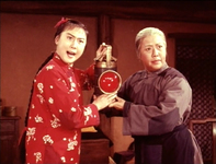 Scene depicting two female characters at center, holding a red signal lantern. On the left is Li Tiemei, with her hair in a long braid and costumed in a red shirt patterned with white flowers. On the right is Granny Li, an elderly woman costumed in a patched blue robe. The background setting is the interior of their home, with simple wooden furniture and beige walls.