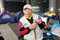 In this photograph, Big Uncle Wong, an elderly man, smiles with his eyes closed. He sits on a chair in the street, wearing a cargo vest and baseball cap and holding a cane with both hands. Behind him, chairs and tents are on the sidewalk in front of a building